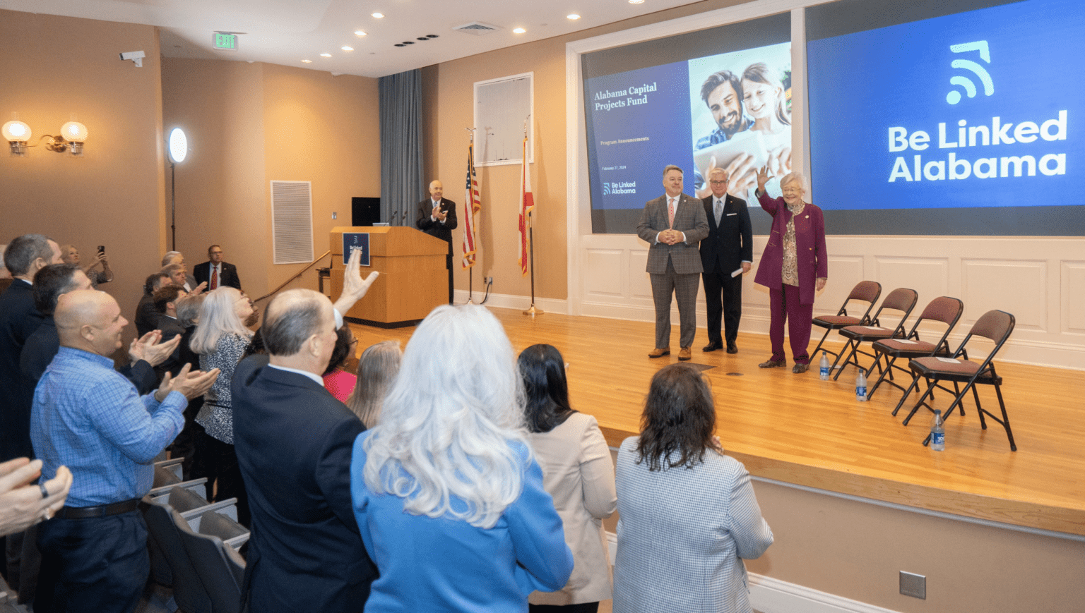 Photograph of Governor Ivey making an announcement with a backdrop displaying the Alabama state flag and the text "Broadband Expansion Initiative.